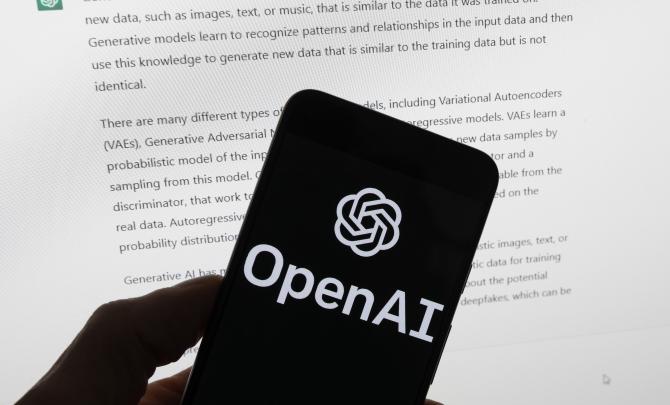 The OpenAI logo is seen on a mobile phone in front of a computer screen which displays output from ChatGPT