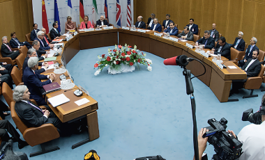 U.S. and Iranian negotiators sit around a cured table as media with cameras and microphones crowd the foreground.