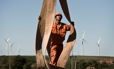 A worker in the foreground adjusts some large straps as he looks up. In the background there is a flat horizon scattered with wind turbines against a blue sky.